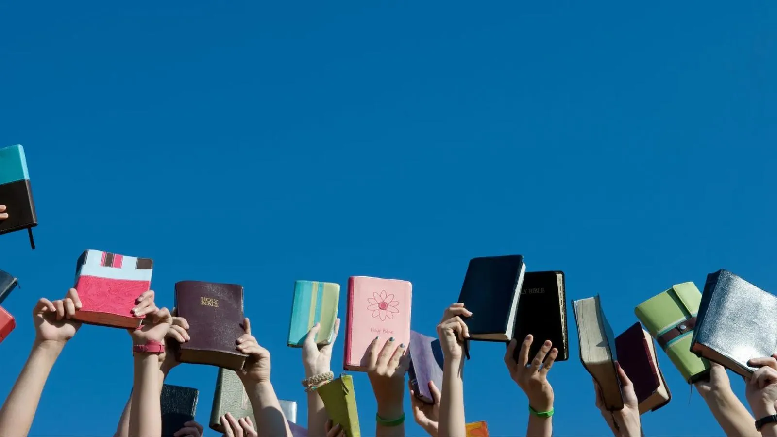 Hands holding up small Bibles against a blue sky
